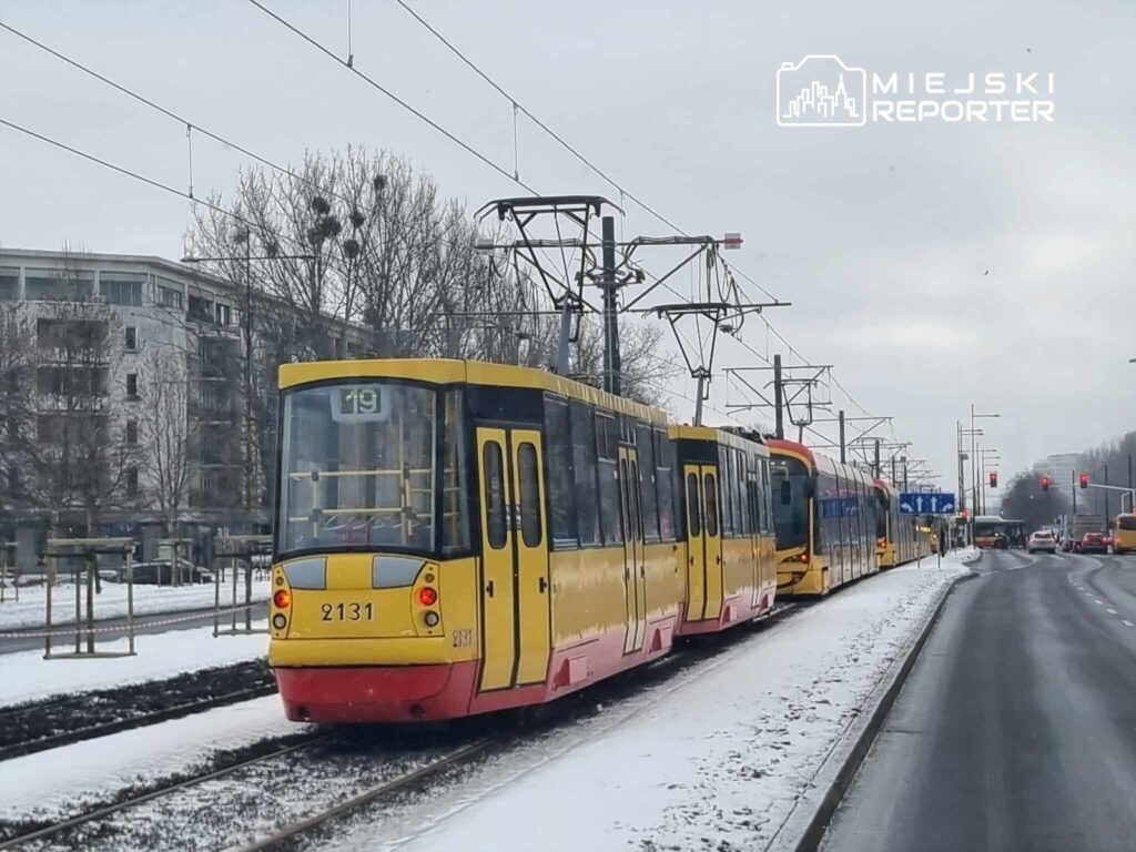 Tramwaj oznaczony numerem 19 zatrzymany na przystanku w zimowej scenerii, obok ruchliwej ulicy.