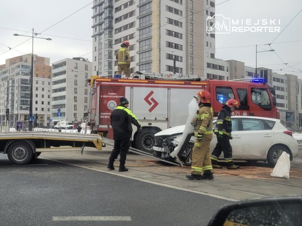 Strażacy w kaskach i mundurach zabezpieczają miejsce wypadku drogowego, obok stoi wóz strażacki.