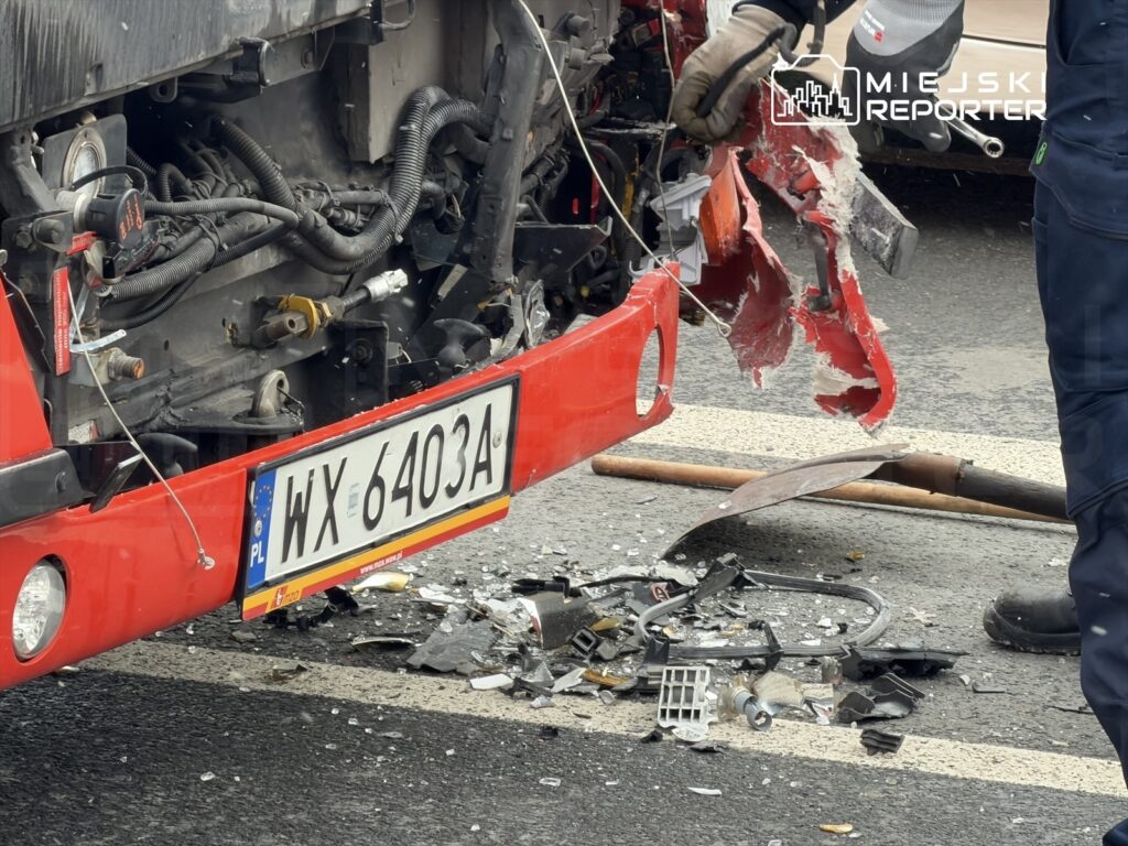 Czerwony autobus z uszkodzoną tylną częścią stoi na drodze, a obok leżą rozbite elementy pojazdu.
