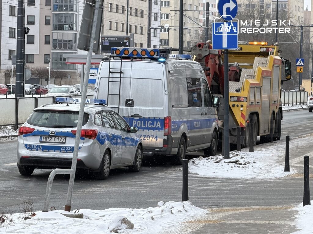 Policyjny radiowóz i furgonetka stoją na jezdni, obok podnośnik służb komunalnych w trakcie pracy. W tle widać nowoczesne budynki.
