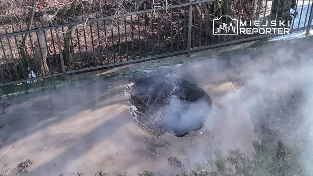 W ziemi powstał duży krater, z którego unosi się dym, otoczony metalowym ogrodzeniem i nagimi drzewami.