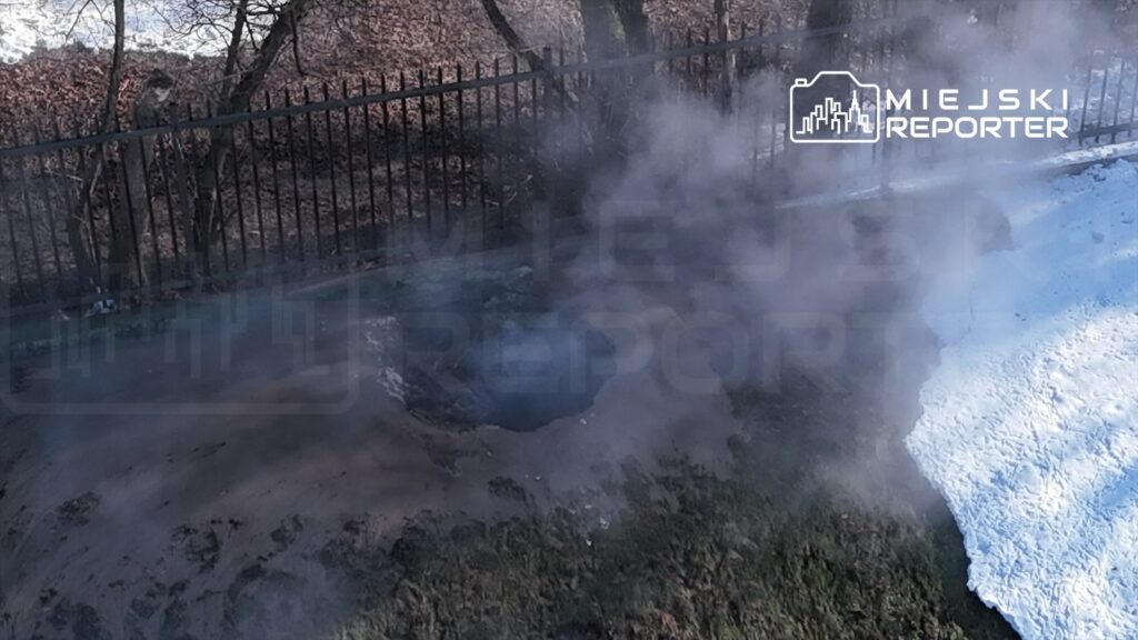 Wydobywająca się para z zagłębienia w ziemi obok ogrodzenia, w otoczeniu nagiej ziemi i resztek śniegu.