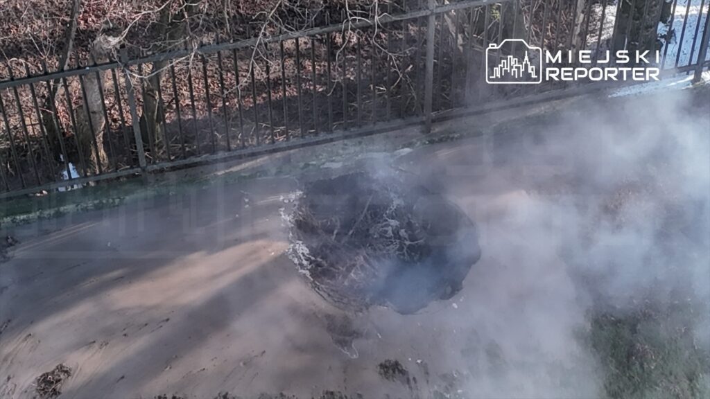 Dym unoszący się z dziury w ziemi, otoczonej metalowym ogrodzeniem, w tle widać nagie drzewa.