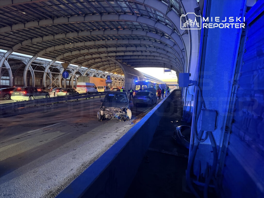 Uszkodzony samochód stoi na pasie ruchu pod zadaszoną konstrukcją, obok zatrzymane pojazdy i pracujący zespół ratunkowy.