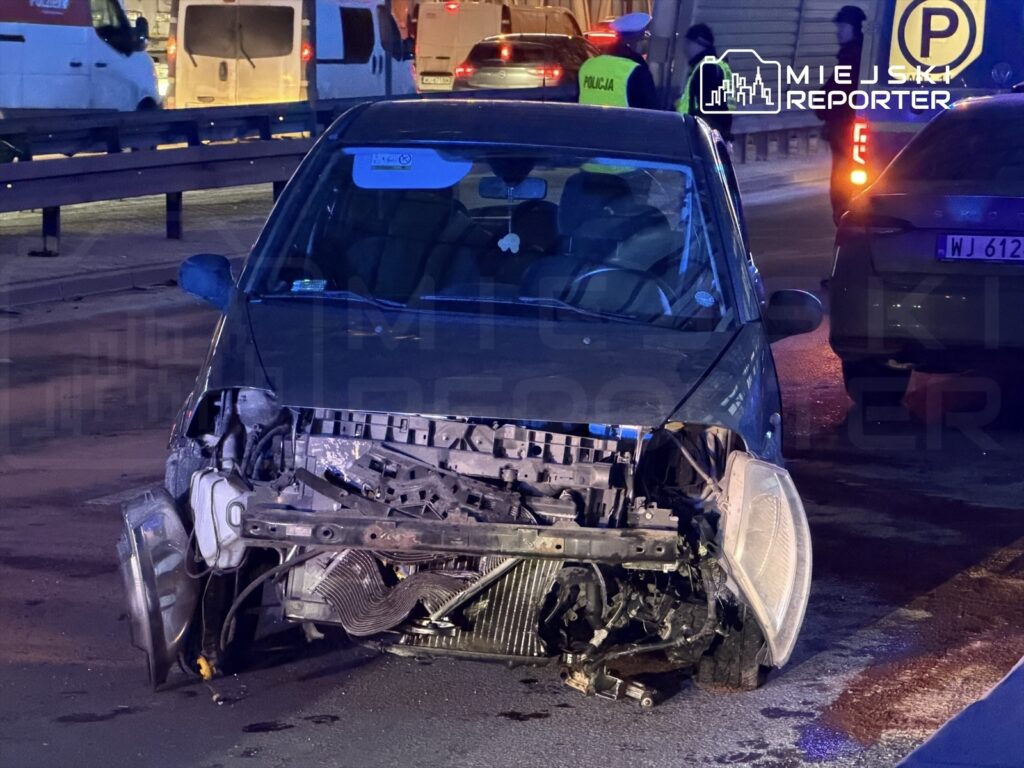 Rozbity samochód osobowy z uszkodzoną przednią częścią stoi na jezdni, obok widać interweniujących policjantów.