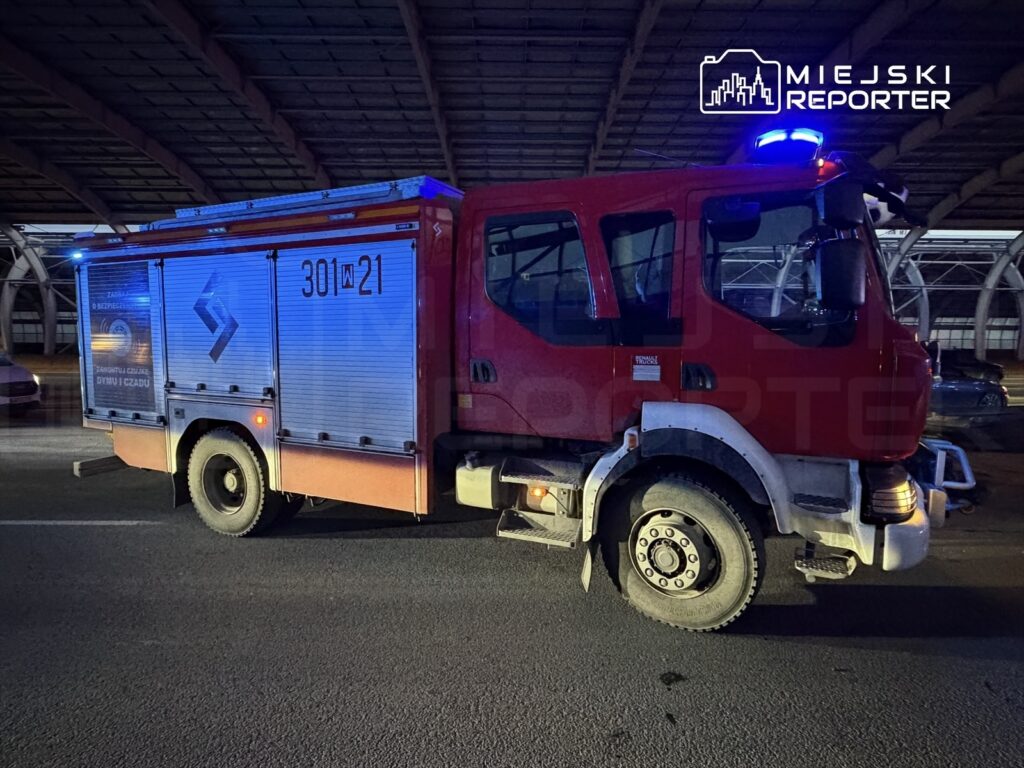 Czerwony wóz strażacki z niebieskimi światłami stojący pod zadaszeniem, na drodze w nocy.