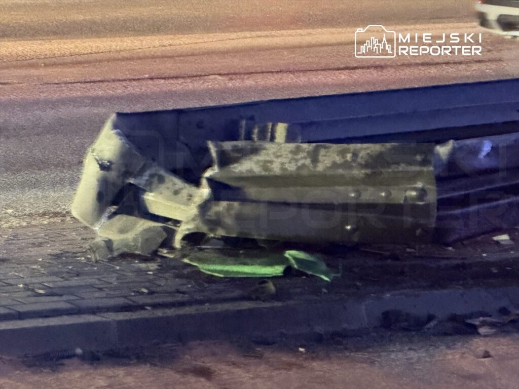 Zniszczony fragment ogrodzenia leży na chodniku obok drogi, w tle widoczne rozmyte światła uliczne.