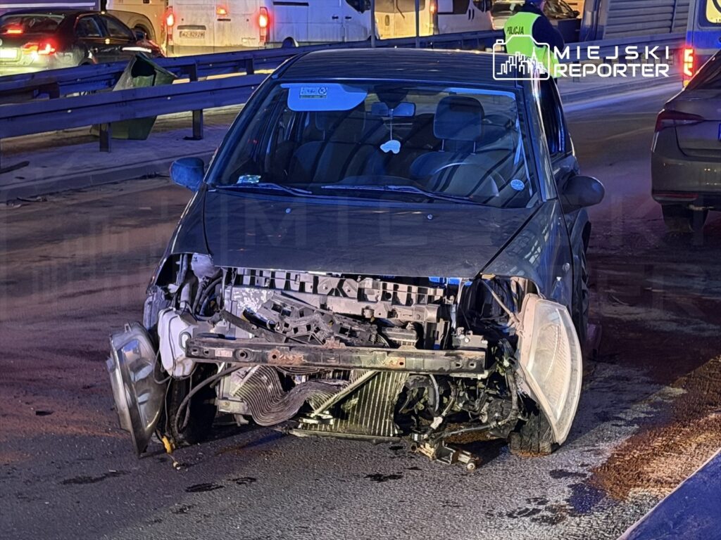 Uszkodzony samochód osobowy z odsłoniętym przodem stoi na jezdni, obok policjant kierujący ruchem.