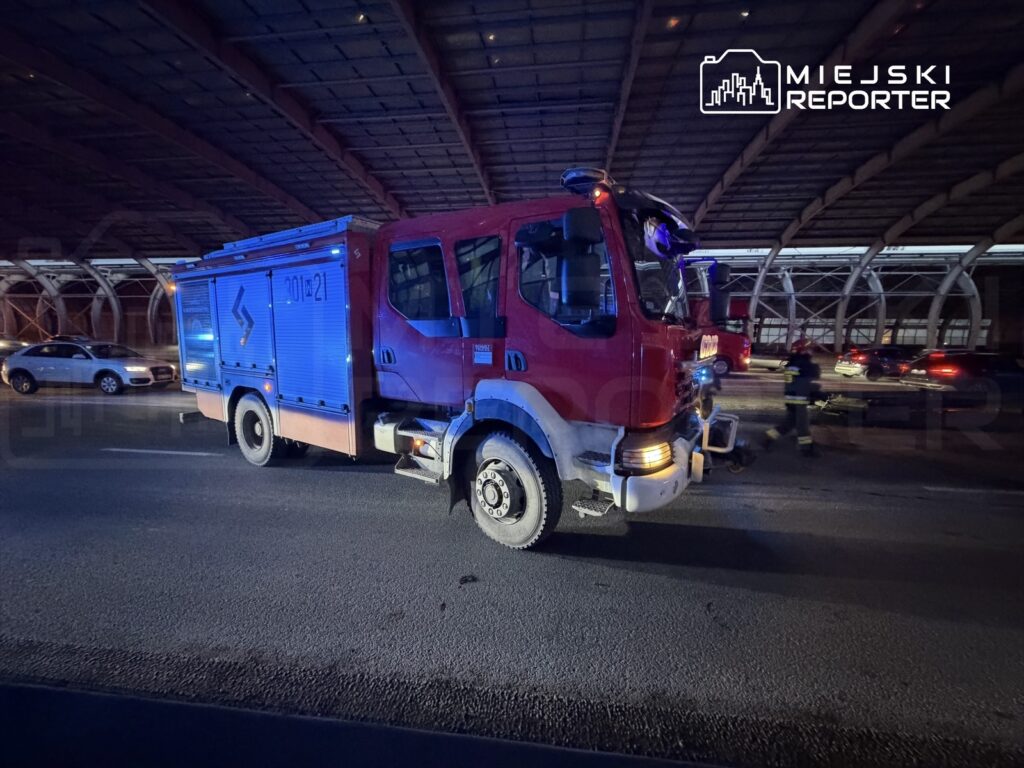 Czerwony wóz strażacki z włączonymi sygnałami świetlnymi zatrzymany na drodze pod zadaszoną konstrukcją.