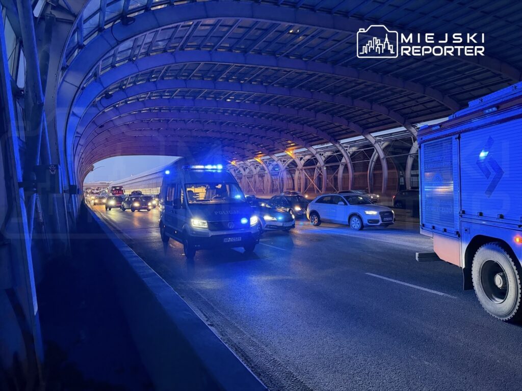 Policyjny radiowóz z niebieskimi światłami przemieszcza się w tunelu, obok stojące samochody w ruchu.