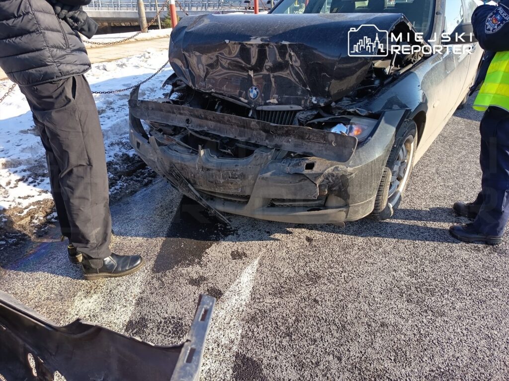 Mężczyzna w czarnej kurtce stoi obok uszkodzonego samochodu osobowego z wgniecionym przodem na zaśnieżonej ulicy.