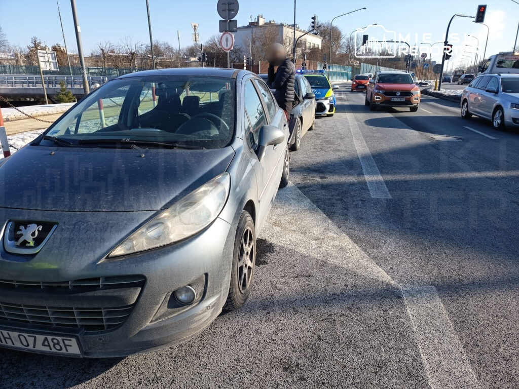 Na ruchliwej ulicy, samochód osobowy marki Peugeot zatrzymał się na poboczu, obok niego stoi mężczyzna w kurtce. W tle widoczny radiowóz policyjny.