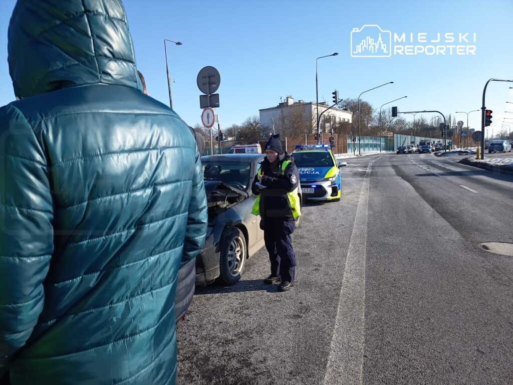 Mężczyzna w puchowej kurtce obserwuje interwencję policji przy uszkodzonym samochodzie na ulicy.