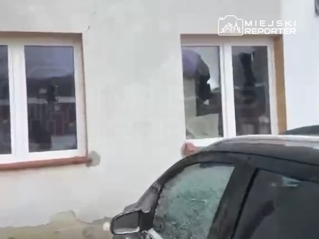 Mężczyzna w ciemnym ubraniu spogląda przez okno budynku, obok zaparkowane samochody.