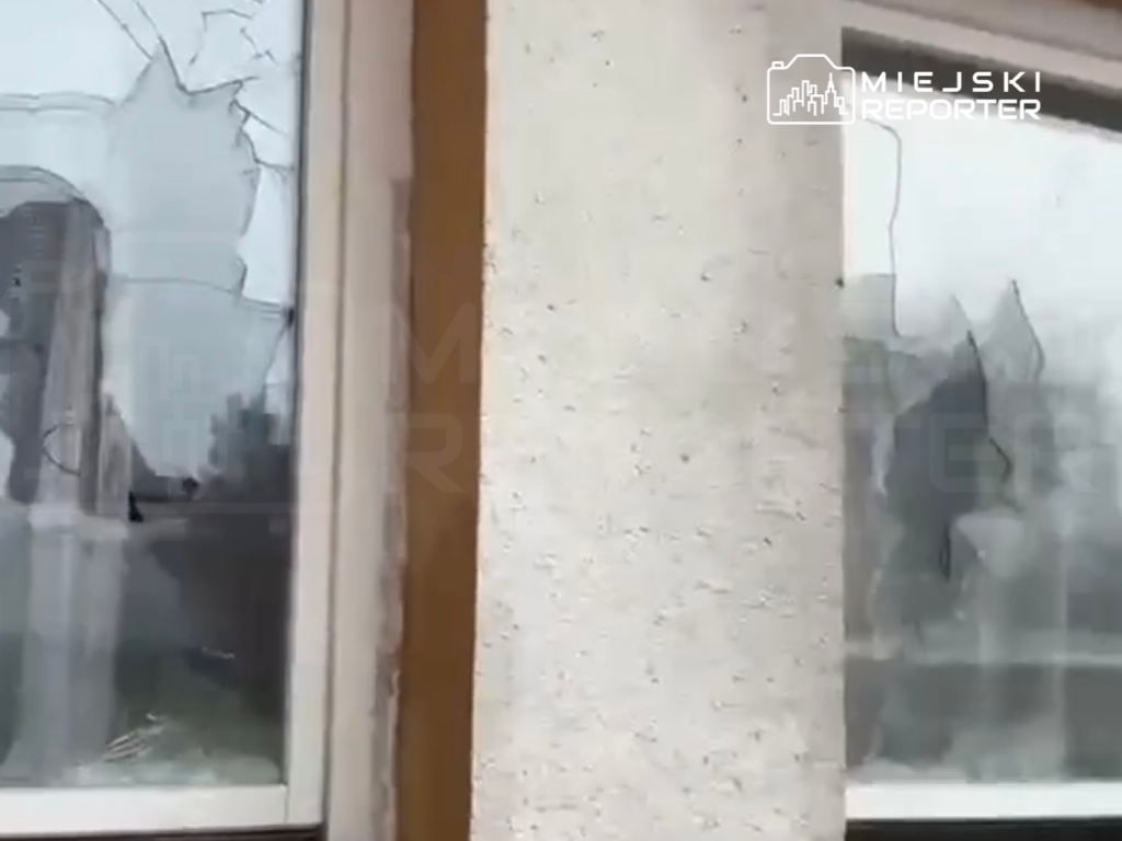 Szyby w oknach z widocznymi pęknięciami i uszkodzeniami, w tle fragmenty budynków.
