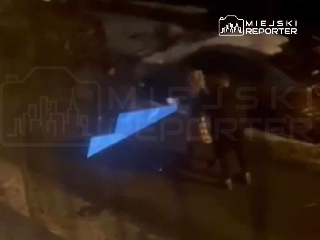 Grupa osób z latarką w ręku znajduje się na ulicy w nocy, oświetlając otoczenie niebieskim światłem.