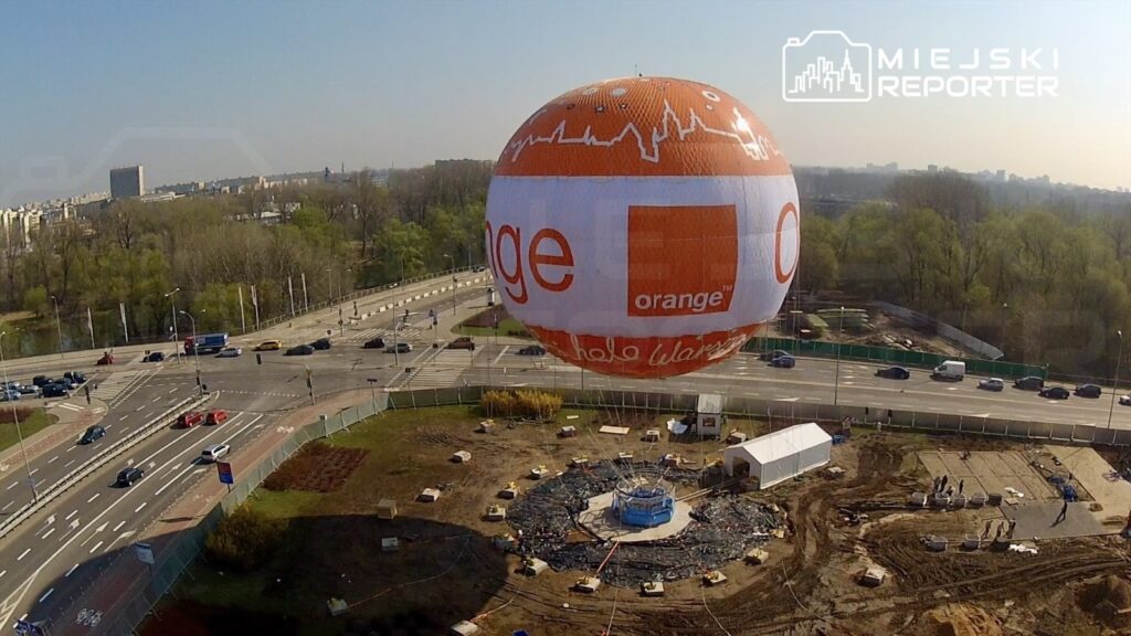 Balon reklamowy w kolorze pomarańczowym unosi się nad placem budowy, w tle widoczne są samochody na ruchliwej ulicy.