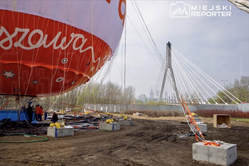 Czerwony balon na gorące powietrze unosi się nad terenem budowy, w tle widać nowoczesny most i skyline miasta.