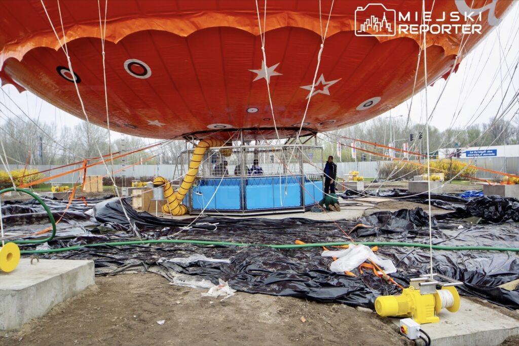 Balon na uwięzi z pomarańczowym kopułą, przygotowany do lotu, otoczony pracownikami oraz sprzętem w budowie.