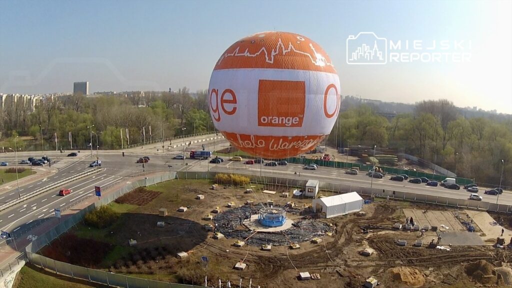 Balon reklamowy w kolorze białym i pomarańczowym unosi się nad placem budowy w miejskim otoczeniu.