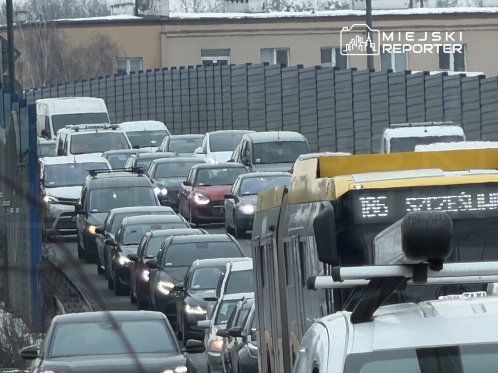 Zatłoczona ulica z wieloma samochodami stojącymi w korku, w tle widoczny autobus z numerem linii.