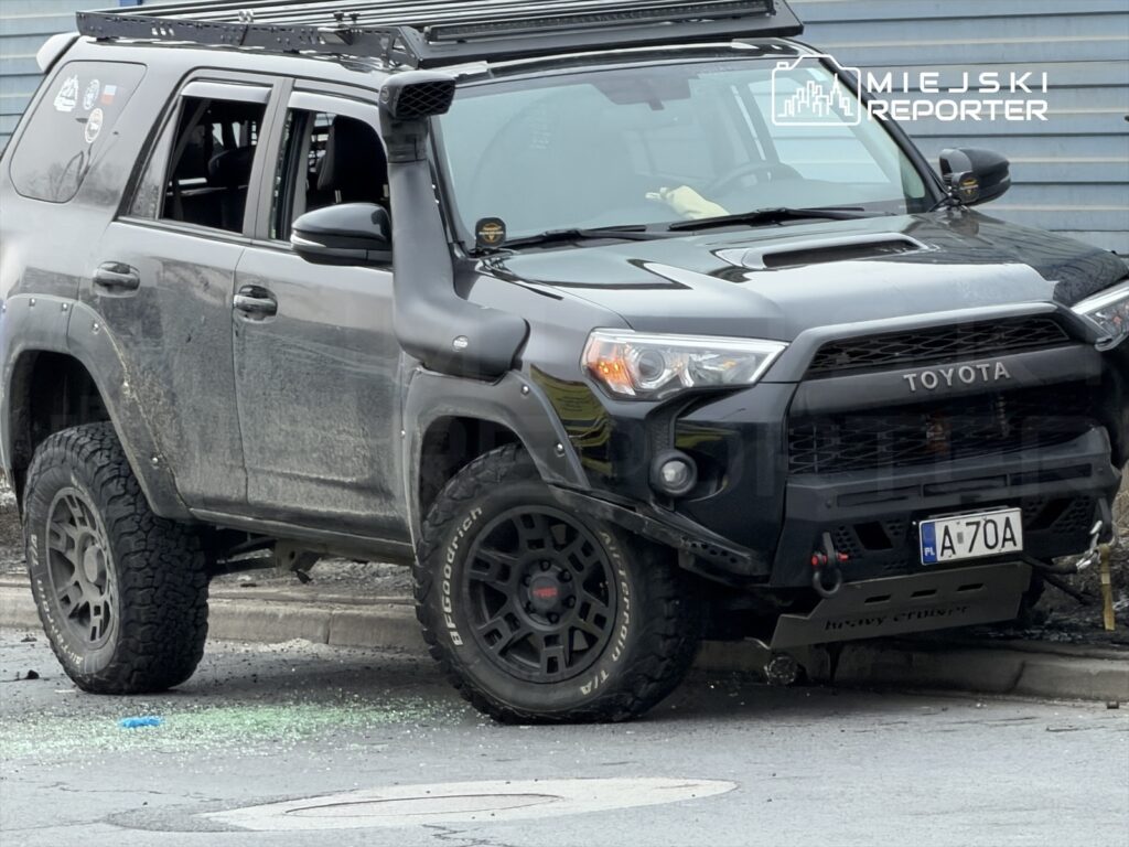 Czarny SUV zaparkowany na krawężniku, z otwartymi oknami i widocznymi śladami uszkodzeń, obok rozbitego szkła.
