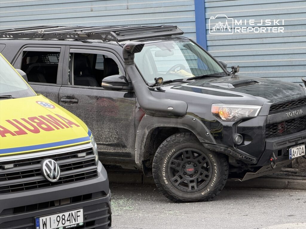 Czarny SUV z modyfikacjami stoi obok żółtej karetki, na tle niebieskiej ściany budynku.
