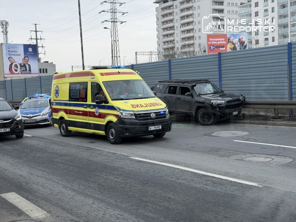 Karetka pogotowia oraz radiowóz policyjny zatrzymane przy uszkodzonym samochodzie na drodze w miejskim otoczeniu.