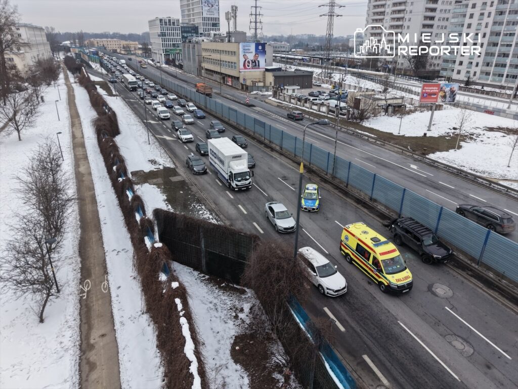 Ruchliwa droga z samochodami osobowymi i ciężarówkami, w tym karetką, obok śnieżnego chodnika i drzew.