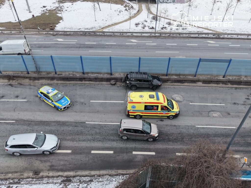 Na drodze poruszają się pojazdy, w tym niebiesko-żółta radiowóz policji oraz żółta karetka pogotowia, obok przejeżdżają inne samochody. W tle widoczne są ogrodzenia i fragmenty śniegu.