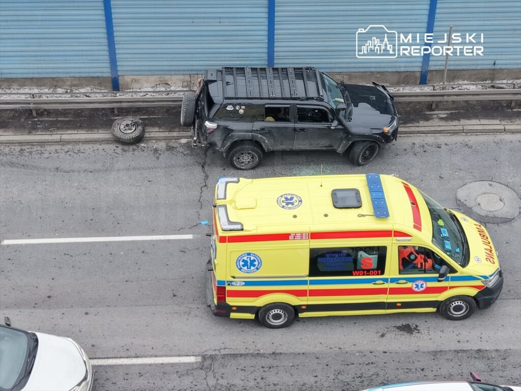 Czarny samochód terenowy z uszkodzoną oponą stoi obok karetki pogotowia na ruchliwej ulicy.
