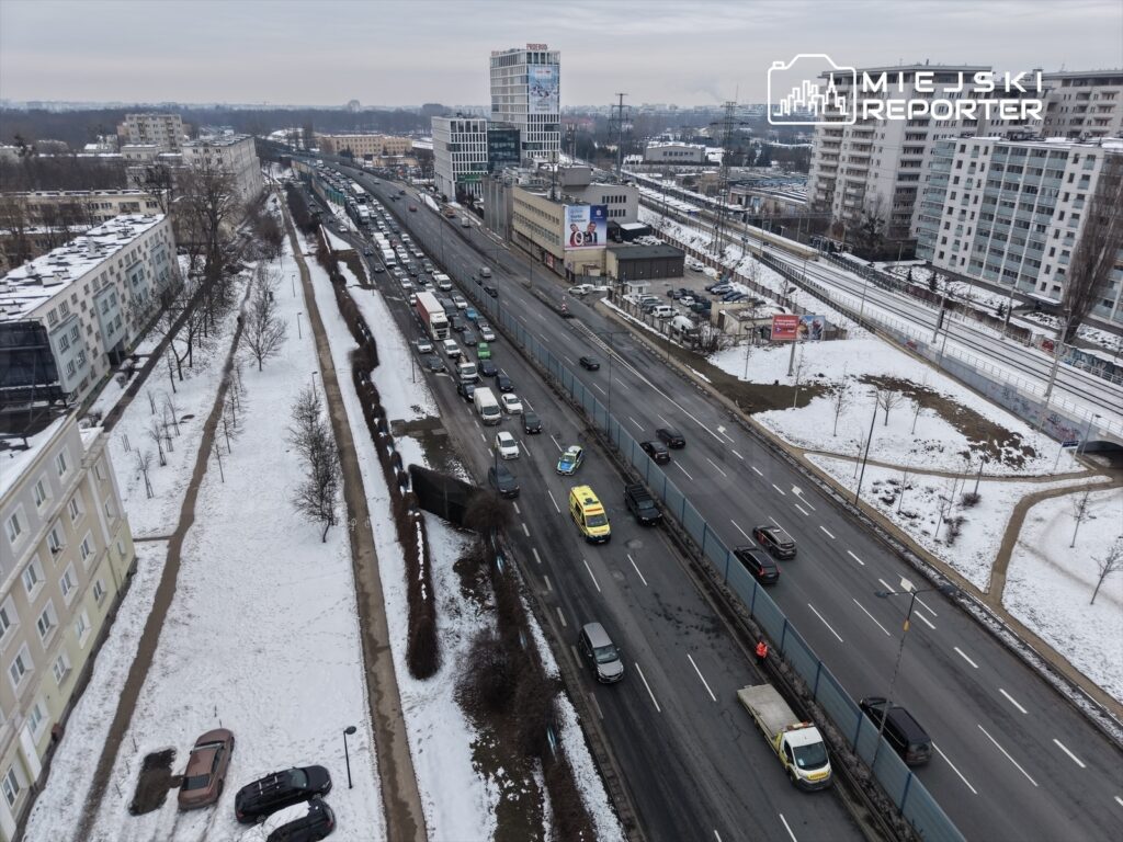 Ruch samochodowy na zatłoczonej drodze, w otoczeniu budynków i pokrytego śniegiem terenu.