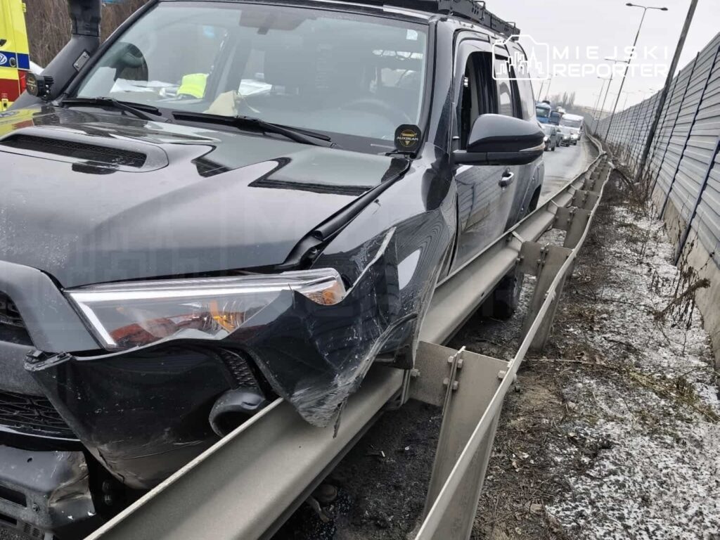 Czarny SUV z uszkodzoną przednią częścią stoi na poboczu drogi, obok bariery ochronnej.
