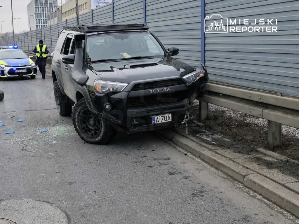 Czarny samochód terenowy zaparkowany przy barierce na ulicy, obok policyjny radiowóz z włączonymi sygnałami świetlnymi.