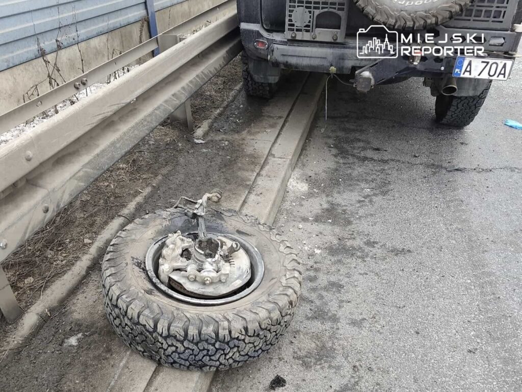 Uszkodzone koło leżące na poboczu drogi obok pojazdu terenowego z widocznym brudem na oponach.