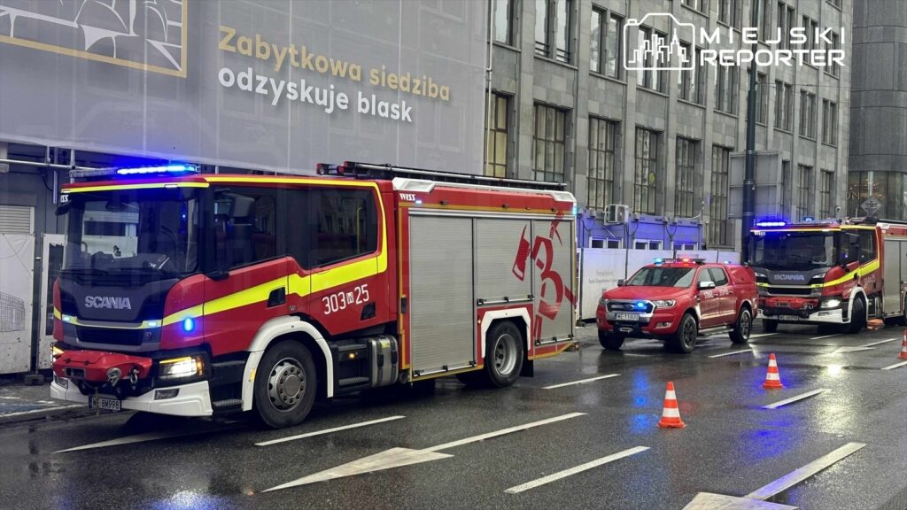 Czerwony wóz strażacki z włączonymi sygnałami świetlnymi stoi na ulicy obok radiowozu policyjnego.