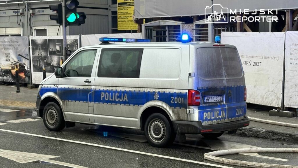 Policyjny van z niebieskimi światłami zatrzymany na ulicy, obok budowy z plakatem informacyjnym.