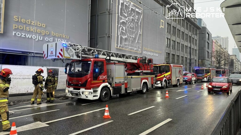 Strażacy w mundurach pracują przy wozach strażackich na ulicy, obok budynku z reklamą banku.