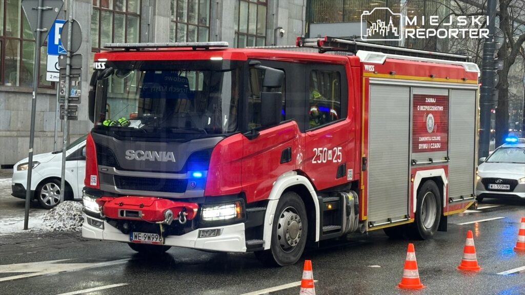 Czerwony wóz strażacki marki Scania zatrzymany na ulicy, obok ustawione pachołki drogowe.