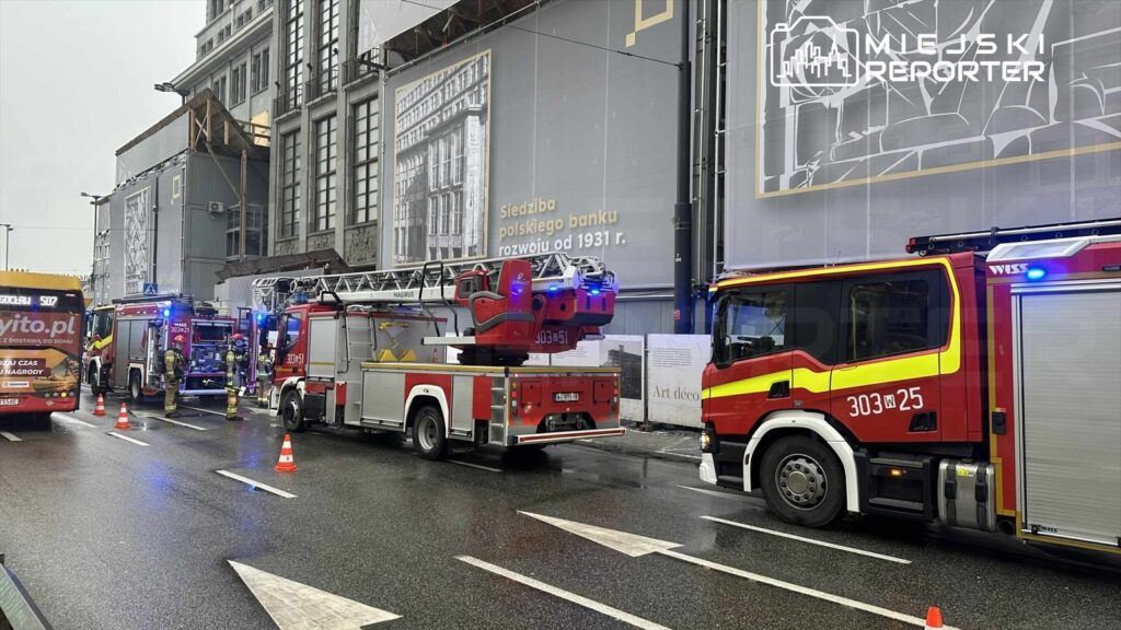Dwa wozy strażackie z włączonymi sygnałami świetlnymi stoją na mokrej nawierzchni, obok budynku w trakcie remontu.
