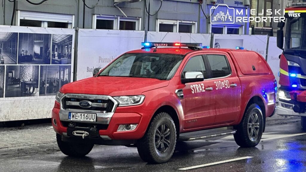 Czerwony samochód strażacki z migającymi światłami zaparkowany na ulicy obok budowy z banerem reklamowym.