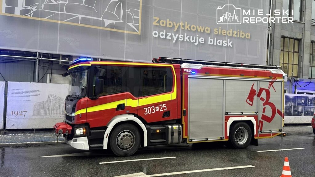 Czerwony wóz strażacki z niebieskimi światłami zatrzymany na ulicy obok placu budowy z reklamą.