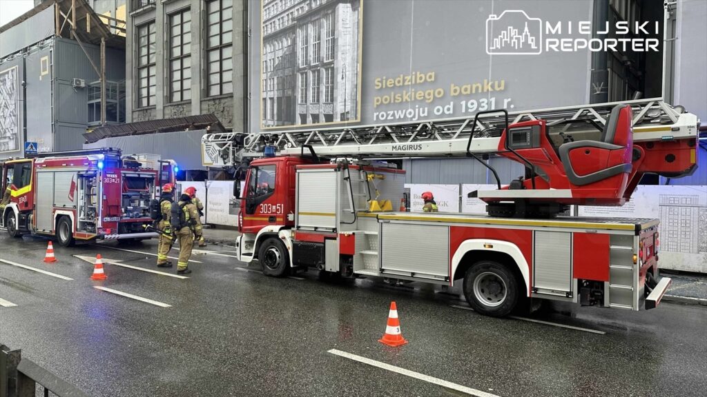 Strażacy w hełmach i kombinezonach pracują obok wozu strażackiego z drabiną, zabezpieczając teren przy budynku.