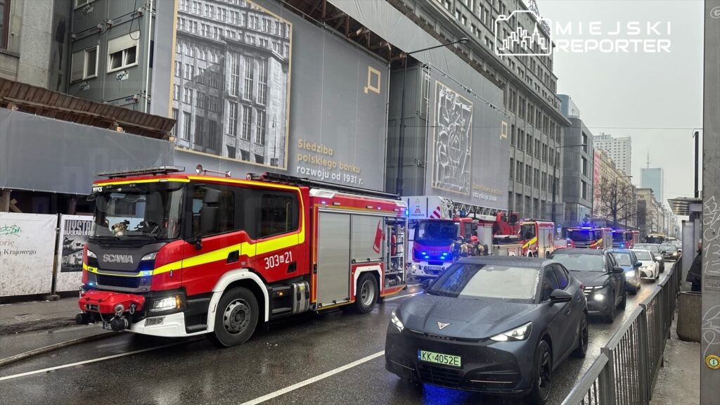 Wielka akcja ratunkowa z udziałem wozów strażackich na ruchliwej ulicy, w tle budynki i reklamy.
