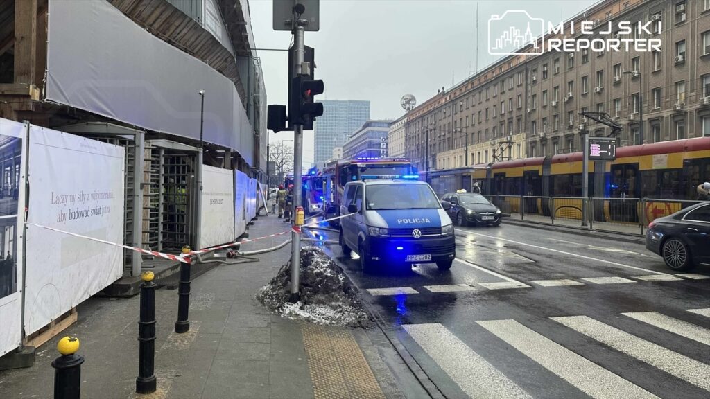 Policyjny radiowóz z włączonymi sygnałami świetlnymi stoi przy ulicy, obok remontowanego budynku.