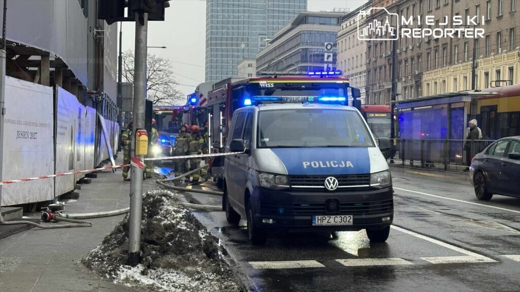 Grupa strażaków pracuje przy wozie strażackim na miejskiej ulicy, obok policyjnego pojazdu z włączonymi sygnałami.