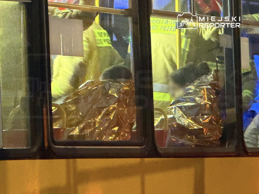 Ratownicy w odzieży ochronnej udzielają pomocy osobom owiniętym w folie termiczne wewnątrz autobusu.
