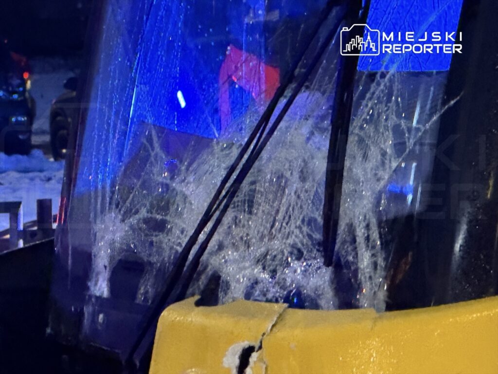 Zniszczona szyba kabiny maszyny budowlanej z widocznymi pęknięciami, w tle niebieskie światła policyjne.