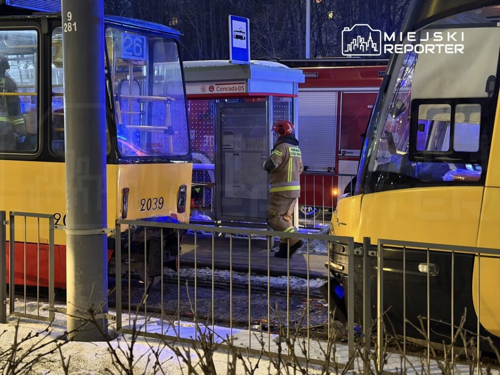 Strażak w hełmie i mundurze zbliża się do tramwaju na przystanku, obok widoczny drugi tramwaj i pojazd strażacki.