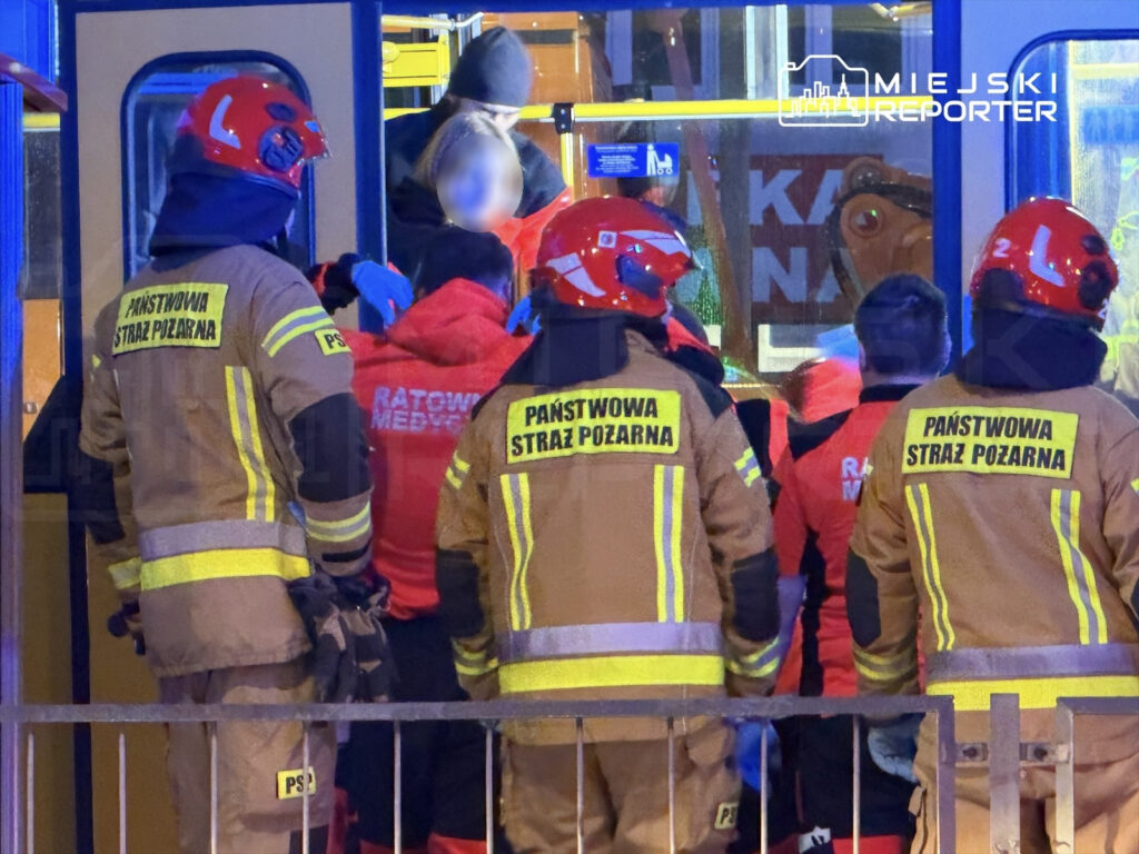 Grupa strażaków w hełmach wspiera ratowników medycznych przy ewakuacji osób z pojazdu w nocy.